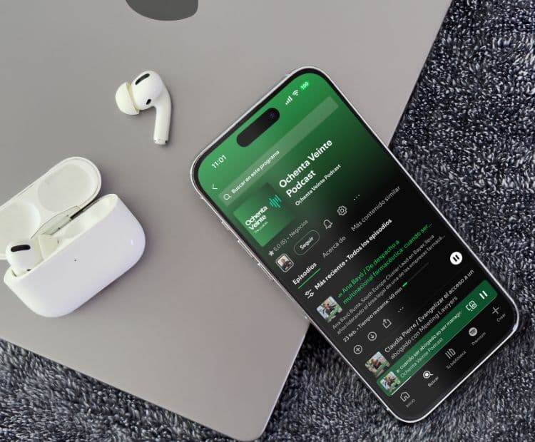 iPhone mostrando Ochenta Veinte Podcast en Spotify junto a unos AirPods y un MacBook, representando el consumo de podcasts legales.